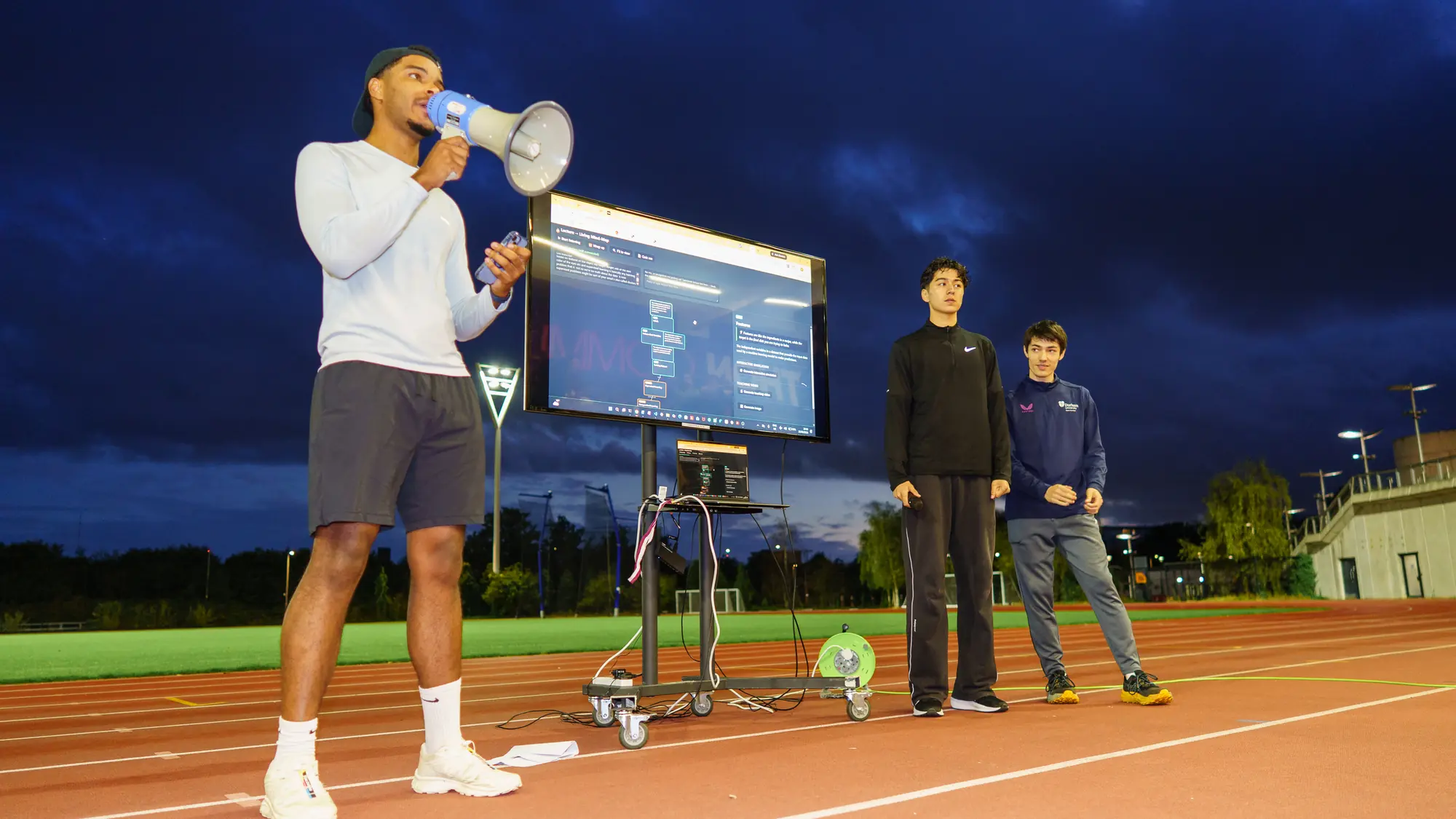 Pitching the project through a megaphone on an athletics track at dusk, with the mind-map displayed on a screen wheeled out beside the lane and two teammates standing to the right.