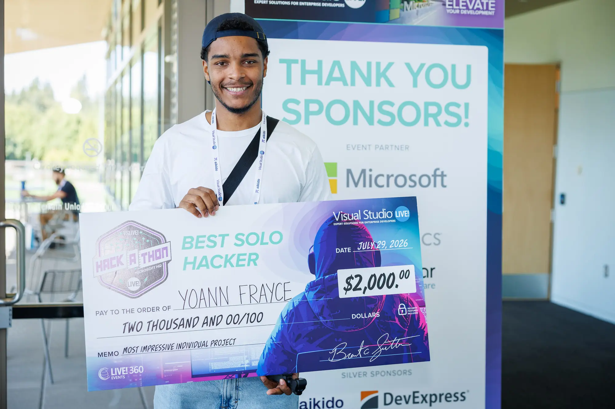 Holding the VSLive! Hack-a-thon award cheque at Microsoft HQ. It reads Best Solo Hacker, made out to Yoann Frayce, dated 29 July 2026, with the memo line “most impressive individual project”. A sponsor banner behind names Microsoft as event partner.