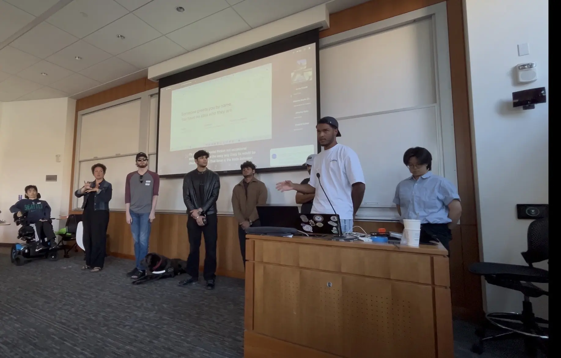 Presenting Omnivision in a lecture theatre at the CREATE Accessibility Hackathon. The screen behind reads “Someone greets you by name. You have no idea who they are.” An ASL interpreter signs beside the stage, and the audience includes a wheelchair user and a guide dog.
